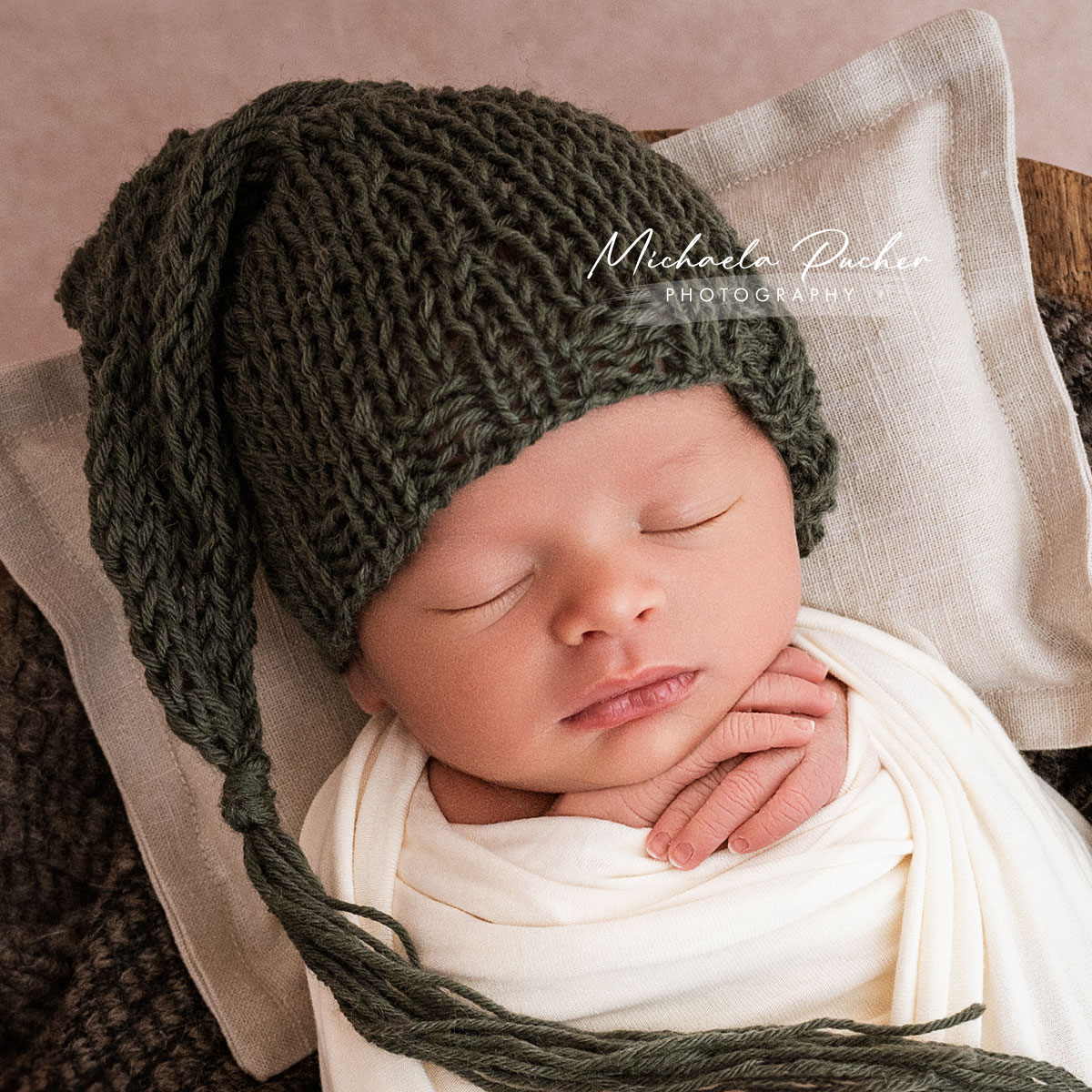 Schlafendes Neugeborenes mit grüner Strickmütze liegt eingewickelt in cremefarbenen Tüchern auf einem kleinen Kissen.